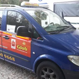 Niebieski minibus Mercedes-Benz z reklamą korporacji taksówkarskiej Glob Taxi w Warszawie, z widocznymi numerami telefonów i logotypem, zaparkowany obok białego kampera.