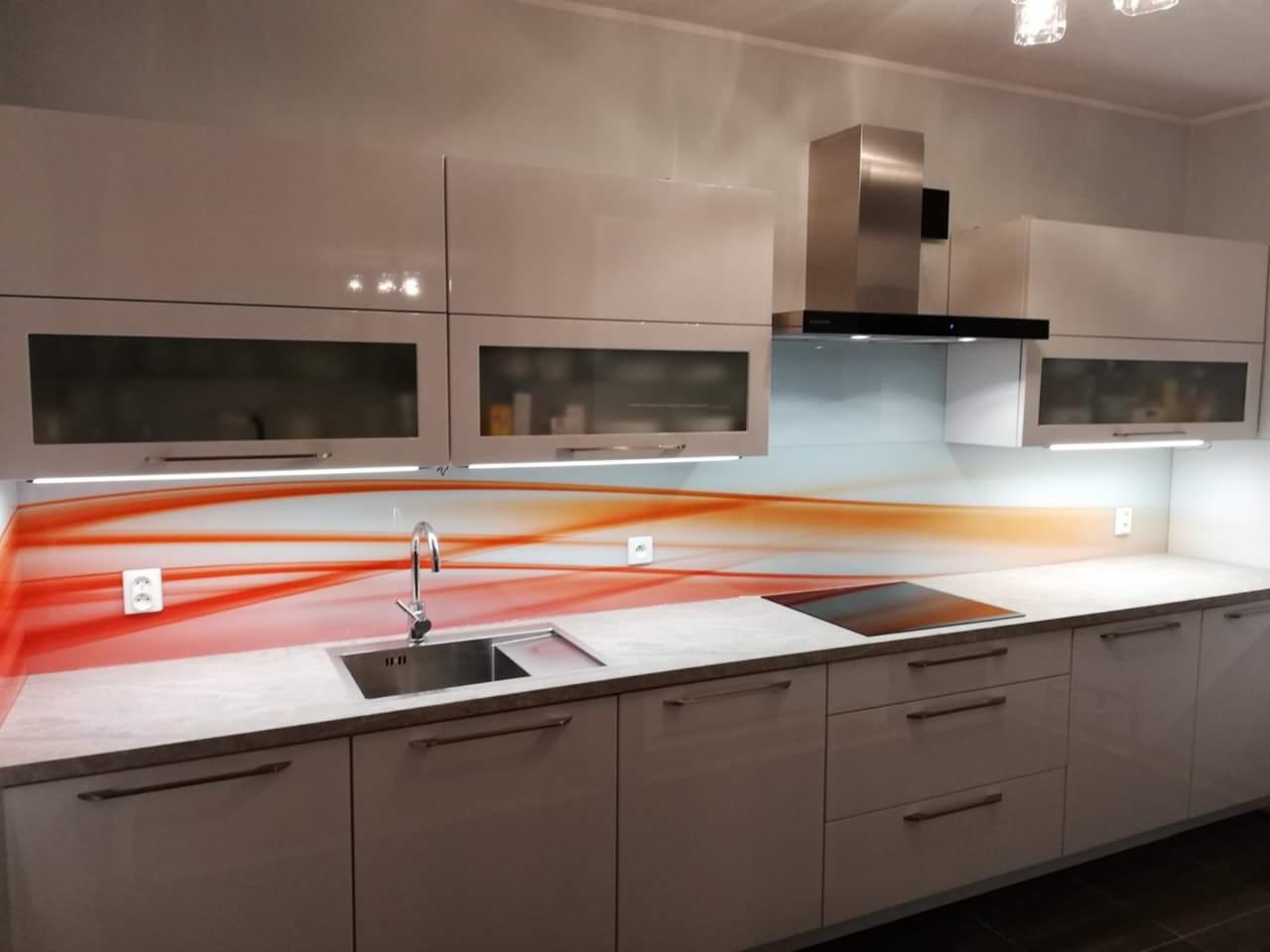 Nowoczesna kuchnia z białymi, błyszczącymi szafkami, pomarańczowo-białym panelem szklanym nad blatem i srebrnym okapem.