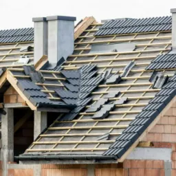 Dom w trakcie budowy, widoczny częściowo położony ciemnoszary dach z dachówki ceramicznej, drewniana konstrukcja więźby dachowej, kominy w stanie surowym, ściany z czerwonej cegły.