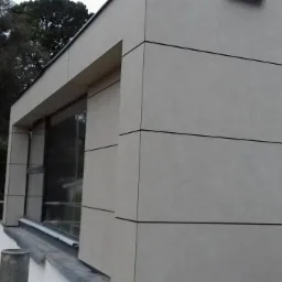 Elewacja budynku wykończona panelami w odcieniach szarości z czarnymi fugami, widoczne rusztowania i zieleń w tle.