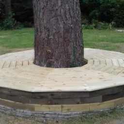 Okrągła drewniana konstrukcja tarasu wokół pnia drzewa na trawniku, widoczne deski ułożone w geometryczny wzór i przewód elektryczny leżący na ziemi.