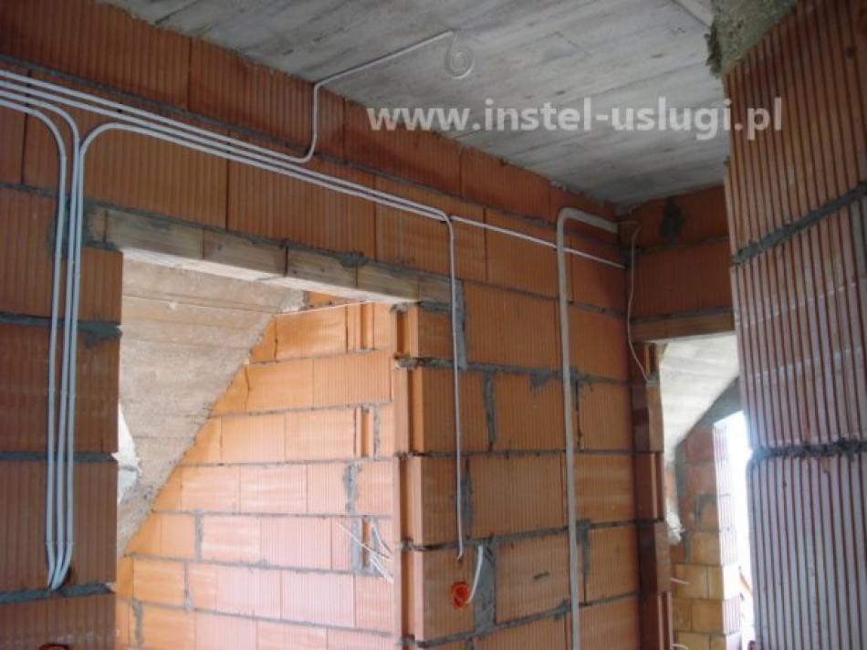 Wnętrze w trakcie budowy: ściany z czerwonej cegły, widoczne białe kable elektryczne prowadzone po ścianach i suficie, otwór drzwiowy w stanie surowym, adres strony internetowej widoczny na górze...