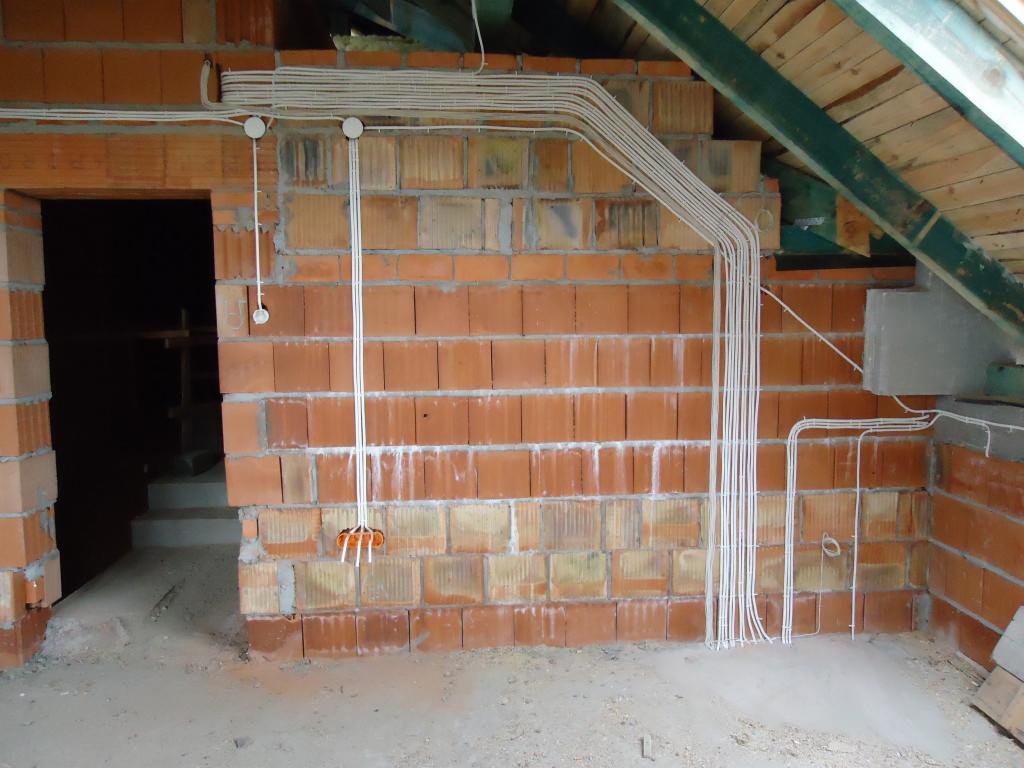 Instalacja elektryczna natynkowa, białe przewody prowadzone po ceglanej ścianie w surowym stanie, widoczne puszki i mocowania, poddasze z drewnianą konstrukcją dachu.