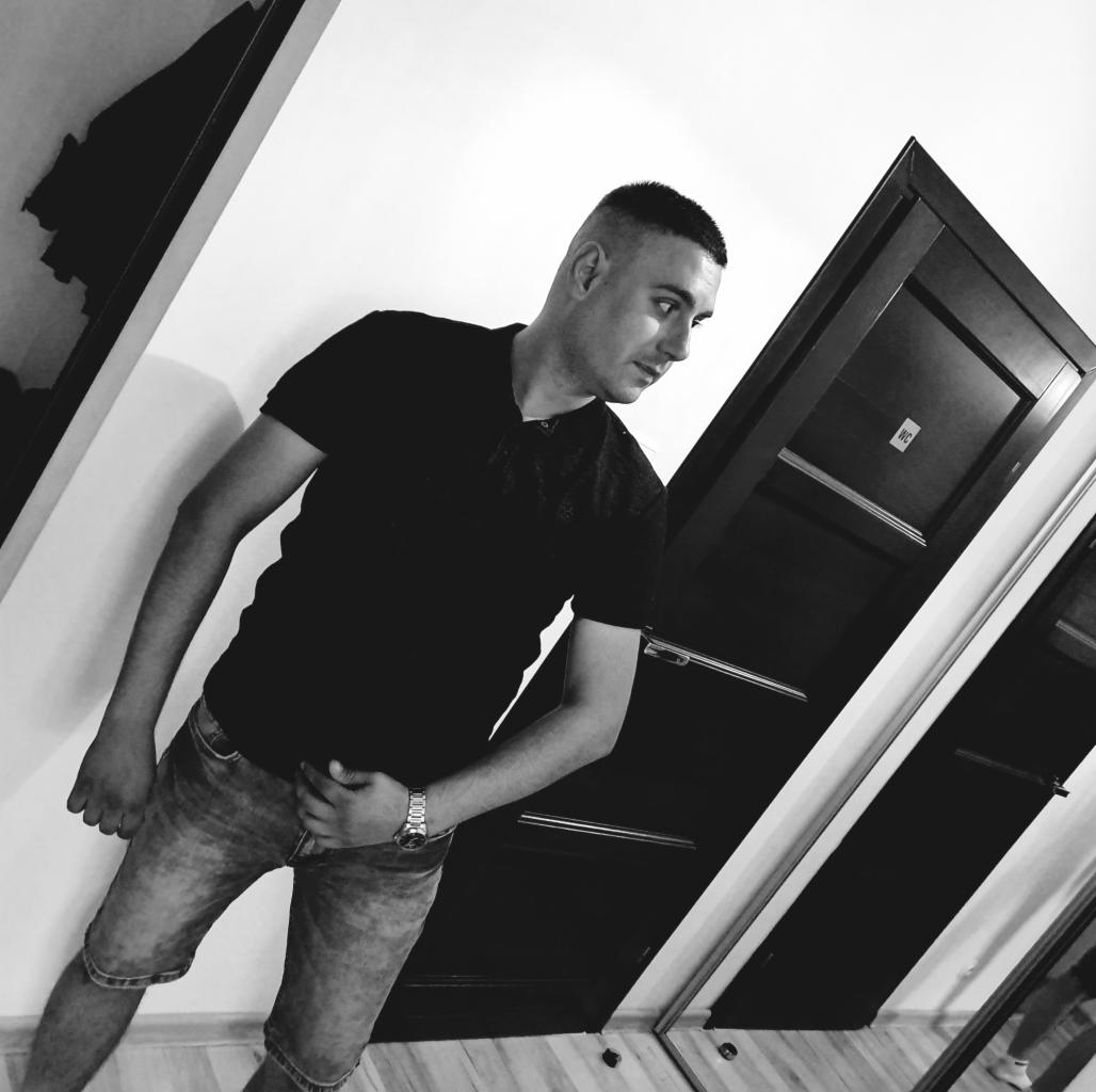 Mężczyzna w czarnej koszulce i jeansowych spodenkach stoi na tle białej ściany i ciemnych drzwi w łazience. Zdjęcie czarno-białe, z nietypowym kątem ujęcia.