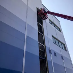 Montaż okna w elewacji budynku przemysłowego z użyciem podnośnika koszowego, widoczna niebieska elewacja panelowa i pracownik w koszu podnośnika.