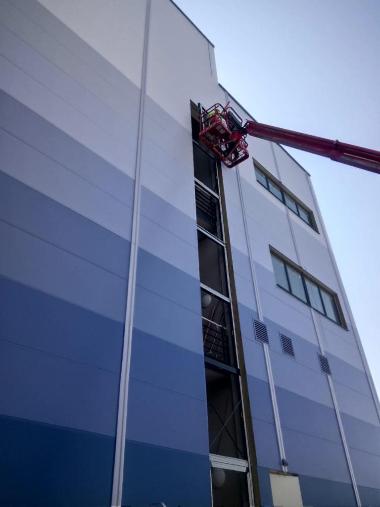 Montaż okna w elewacji budynku przemysłowego z użyciem podnośnika koszowego, widoczna niebieska elewacja panelowa i pracownik w koszu podnośnika.