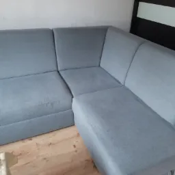 Szara, narożna kanapa z tapicerką o drobnym splocie, ustawiona na drewnianej podłodze, z fragmentem czarnej szafki w tle.