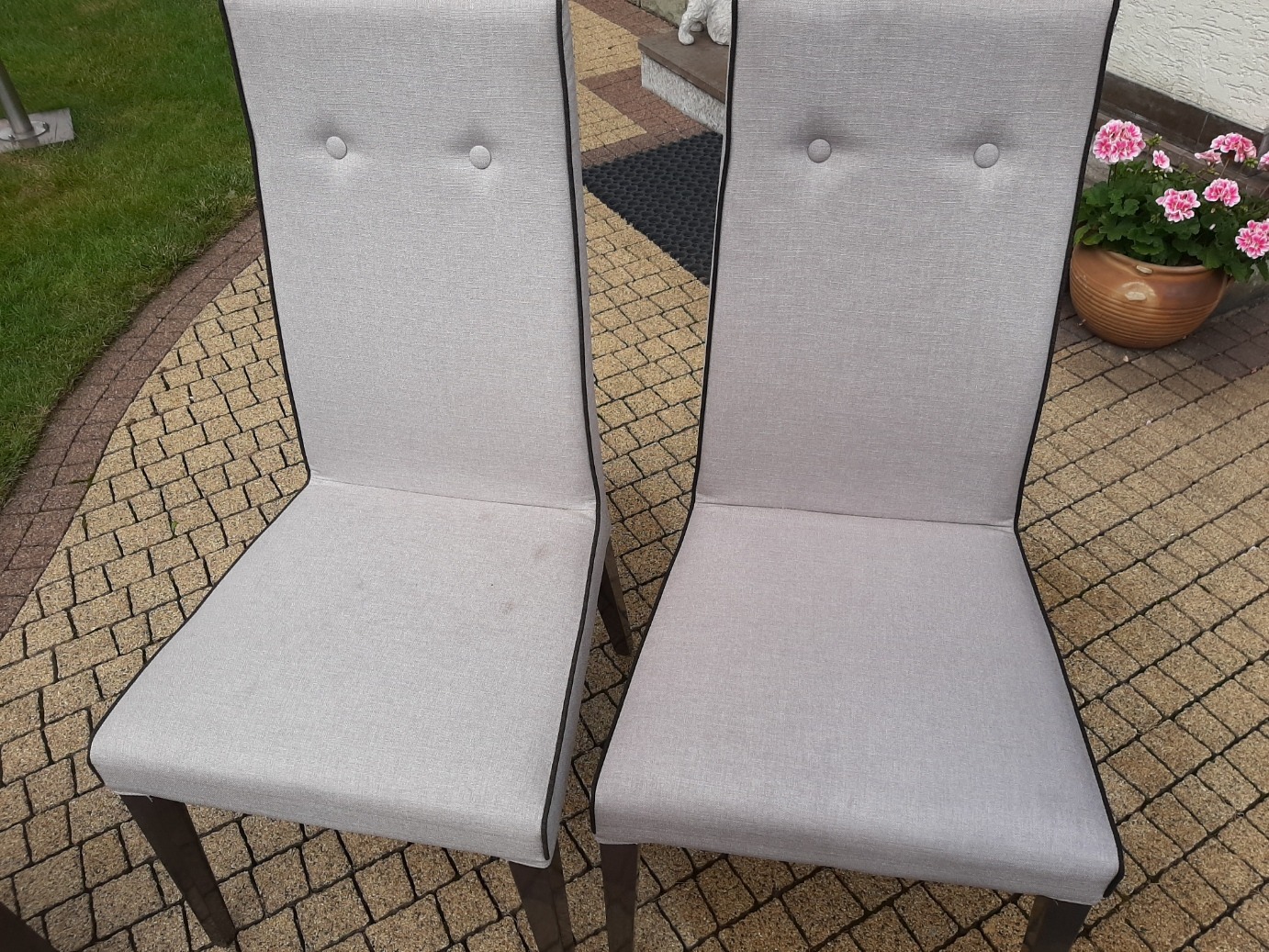 Dwa krzesła tapicerowane jasną tkaniną z ciemną lamówką i dwoma guzikami na oparciu, ustawione na kostce brukowej, jedno z widocznym plamą na siedzisku.