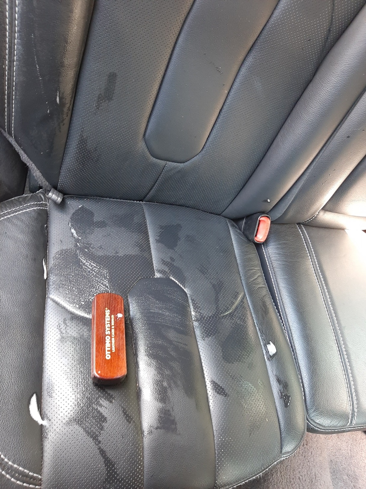 Czyszczone skórzane siedzenie samochodowe z widocznymi plamami i wilgocią, z drewnianym aplikatorem preparatu do czyszczenia skóry marki Ottino Systems. Widoczny fragment pasa bezpieczeństwa...