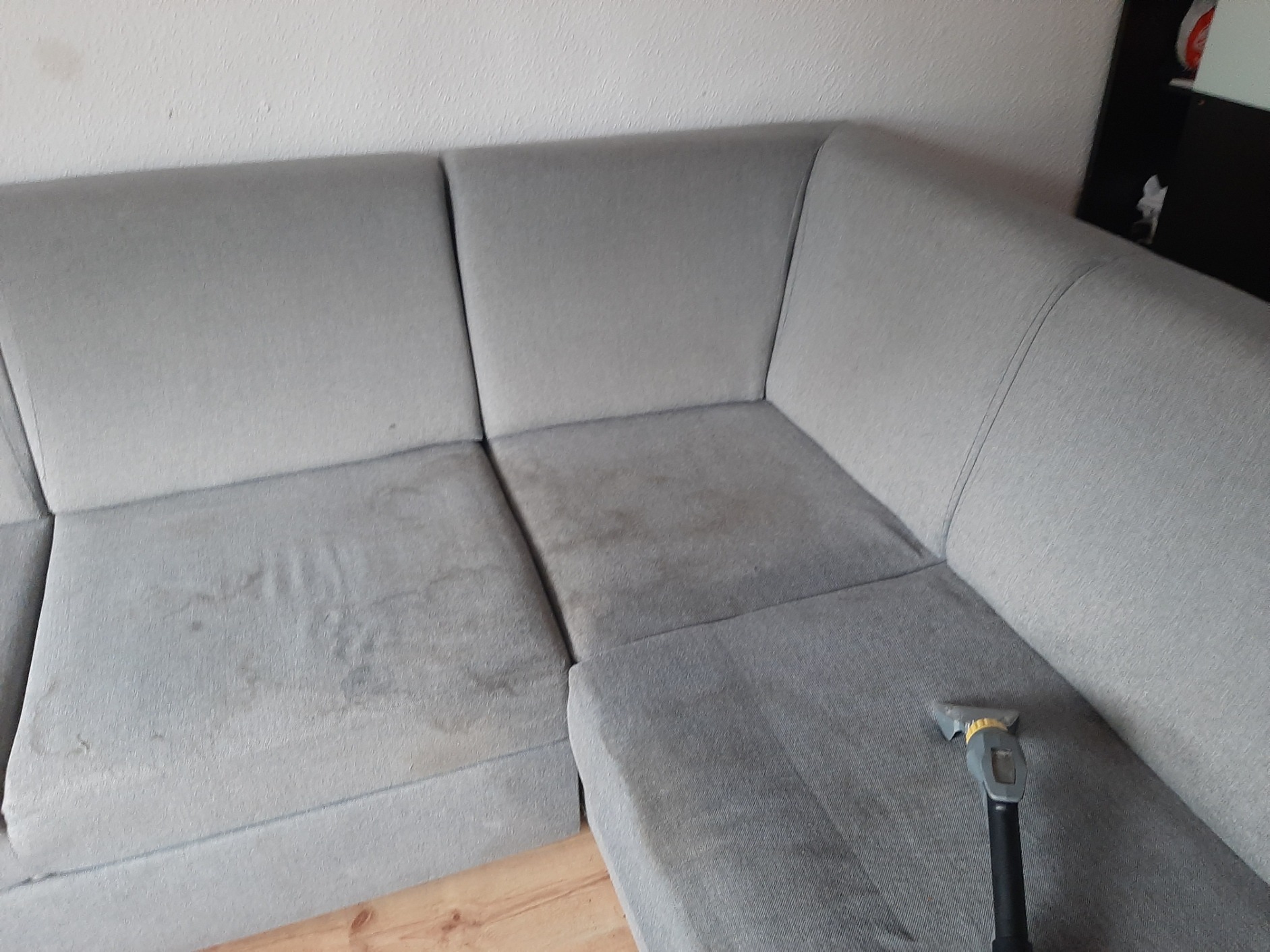 Szara narożna kanapa z widocznymi plamami podczas czyszczenia przy użyciu odkurzacza piorącego z plastikową dyszą.