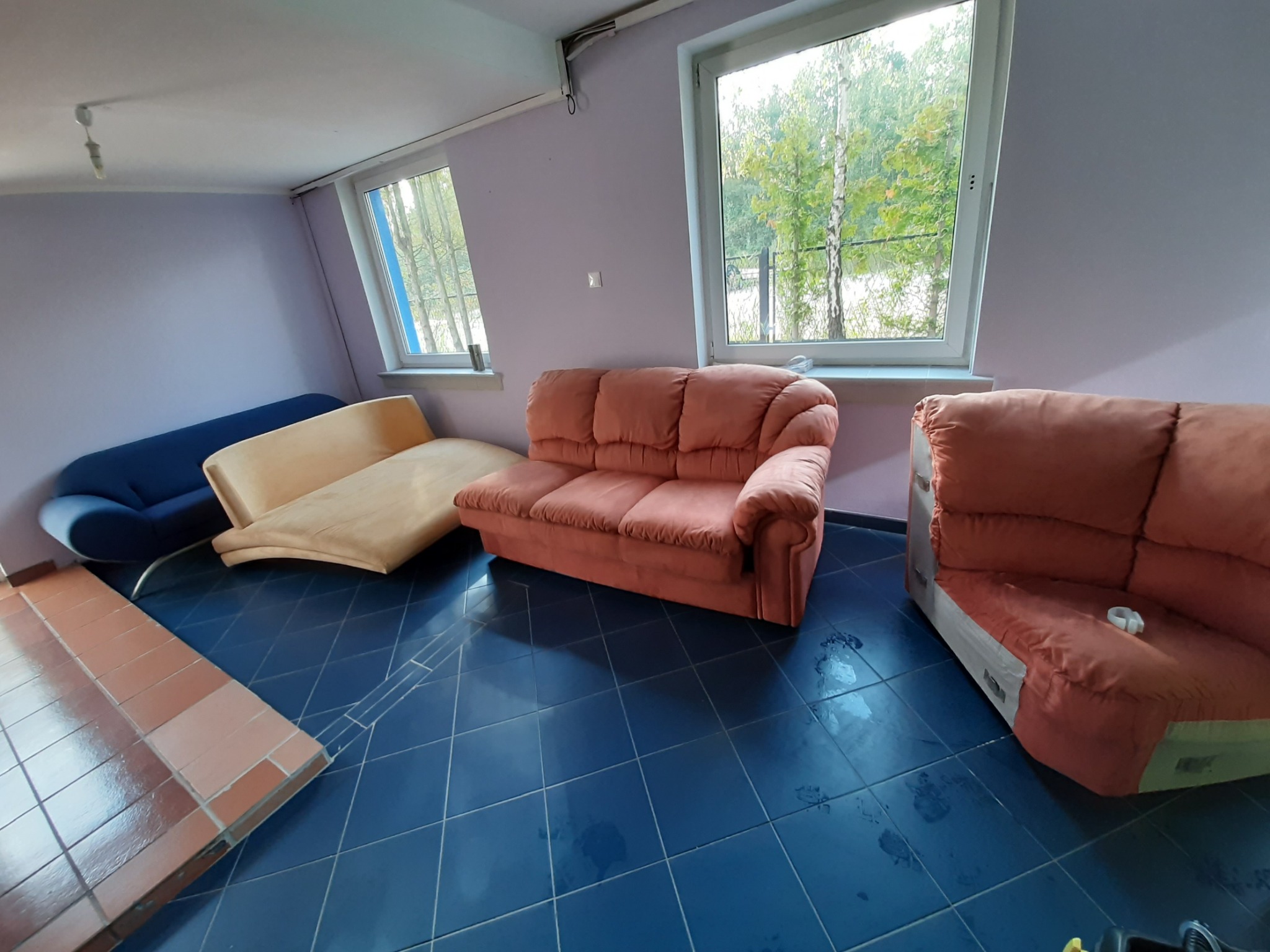 Wnętrze pokoju z trzema sofami o różnych kolorach i kształtach, mokra, niebieska podłoga z płytek, jasne ściany i dwa okna z widokiem na drzewa.