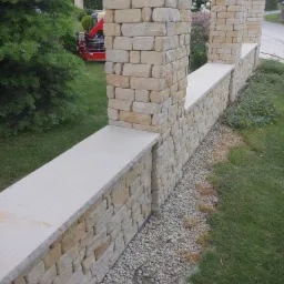 Murowany płot z piaskowca z kamiennymi słupkami w ogrodzie, widoczny czerwony dźwig w tle.