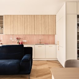 Ambiente Dominika Cymerman - Jasne wnętrze z aneksem kuchennym: różowe płytki nad blatem, drewniane szafki, białe fronty, granatowa sofa i parkiet w jodełkę. Nowoczesny minimalizm.