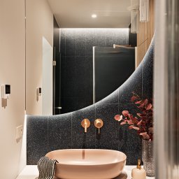 Ambiente Dominika Cymerman - Nowoczesna łazienka z umywalką w odcieniu pudrowego różu, złotą armaturą i lustrem o nieregularnym kształcie. Czarna ściana z mozaiką i drewniane szafki dodają elegancji.