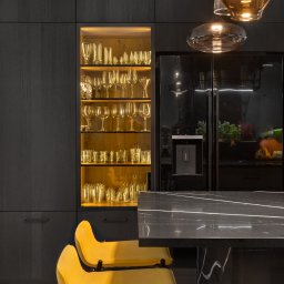 Ambiente Dominika Cymerman - Nowoczesna kuchnia w ciemnych barwach z podświetlaną witryną pełną szkła, czarną lodówką i żółtymi krzesłami barowymi. Lampa wisząca z bursztynowego szkła.