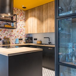 Ambiente Dominika Cymerman - Nowoczesna kuchnia z żółtym sufitem i wzorzystą tapetą. Czarna wyspa z drewnianym blatem, czarne szafki i armatura. W tle przeszklona witryna i geometryczna podłoga.