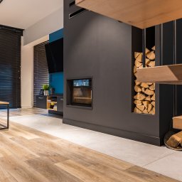 Ambiente Dominika Cymerman - Salon z kominkiem i drewnem opałowym, drewniana podłoga, szare meble, niebieska ściana. Nowoczesne wnętrze z elementami naturalnymi i minimalistycznym designem.