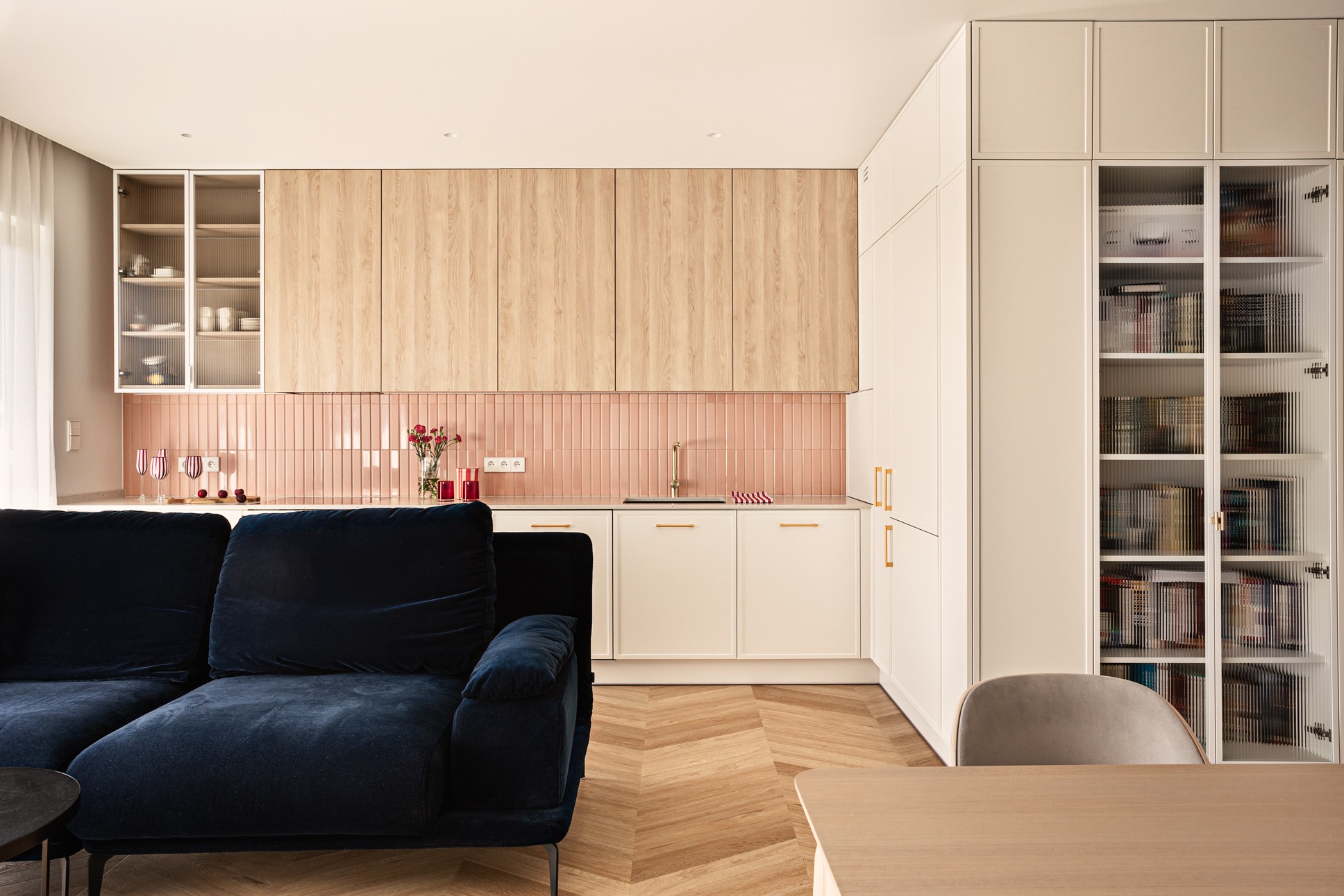 Jasne wnętrze z aneksem kuchennym: różowe płytki nad blatem, drewniane szafki, białe fronty, granatowa sofa i parkiet w jodełkę. Nowoczesny minimalizm.
