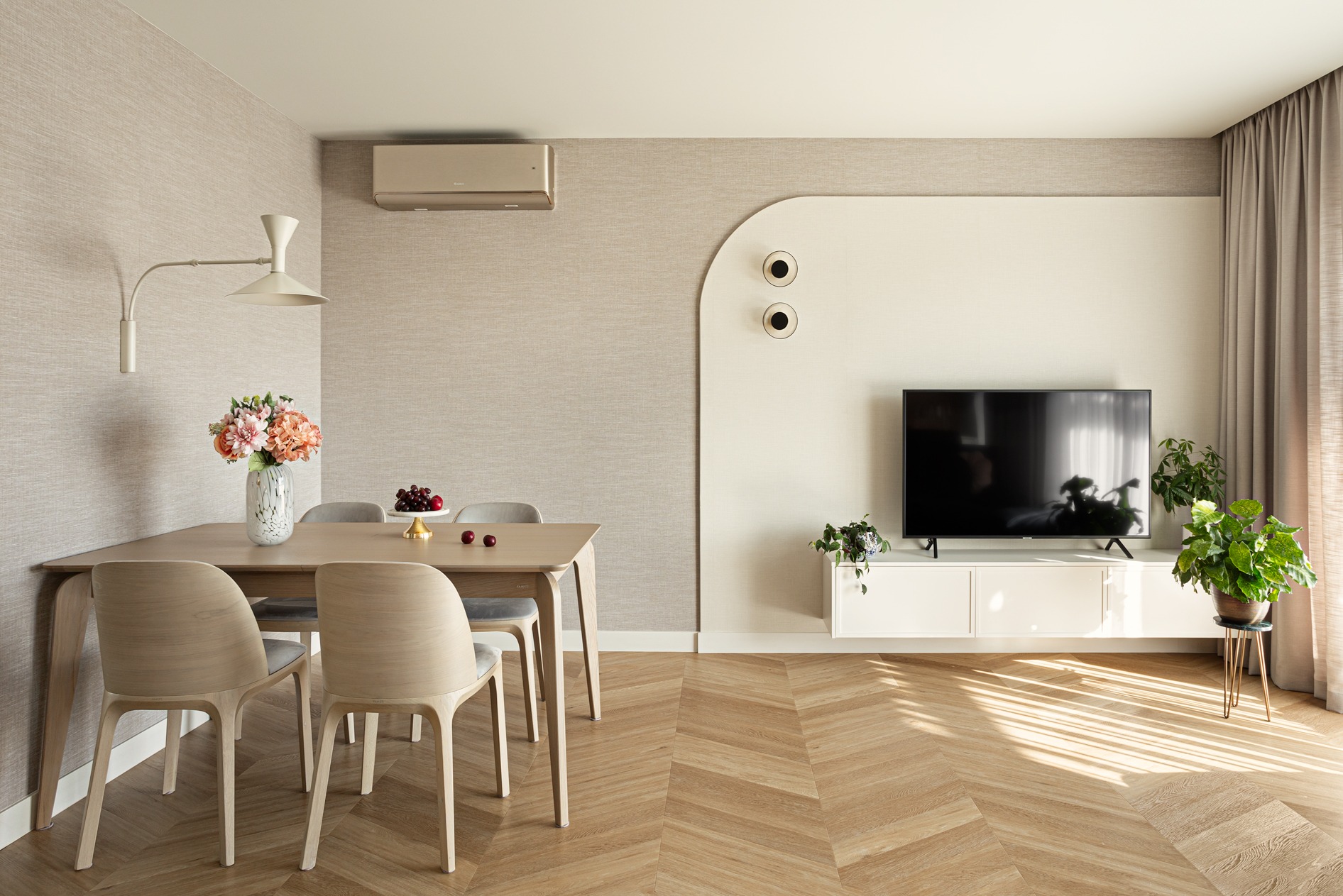 Jasne wnętrze jadalni z parkietem w jodełkę, drewnianym stołem i krzesłami. Na ścianie telewizor i dekoracyjne oświetlenie. Minimalistyczny design z roślinami doniczkowymi.