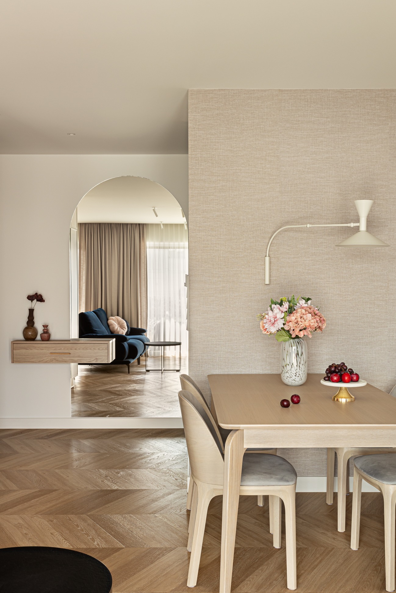 Jasne wnętrze z parkietem w jodełkę, łukowatym przejściem do salonu z granatową sofą, stół z kwiatami i owocami, lampa ścienna. Neutralna kolorystyka, naturalne światło.