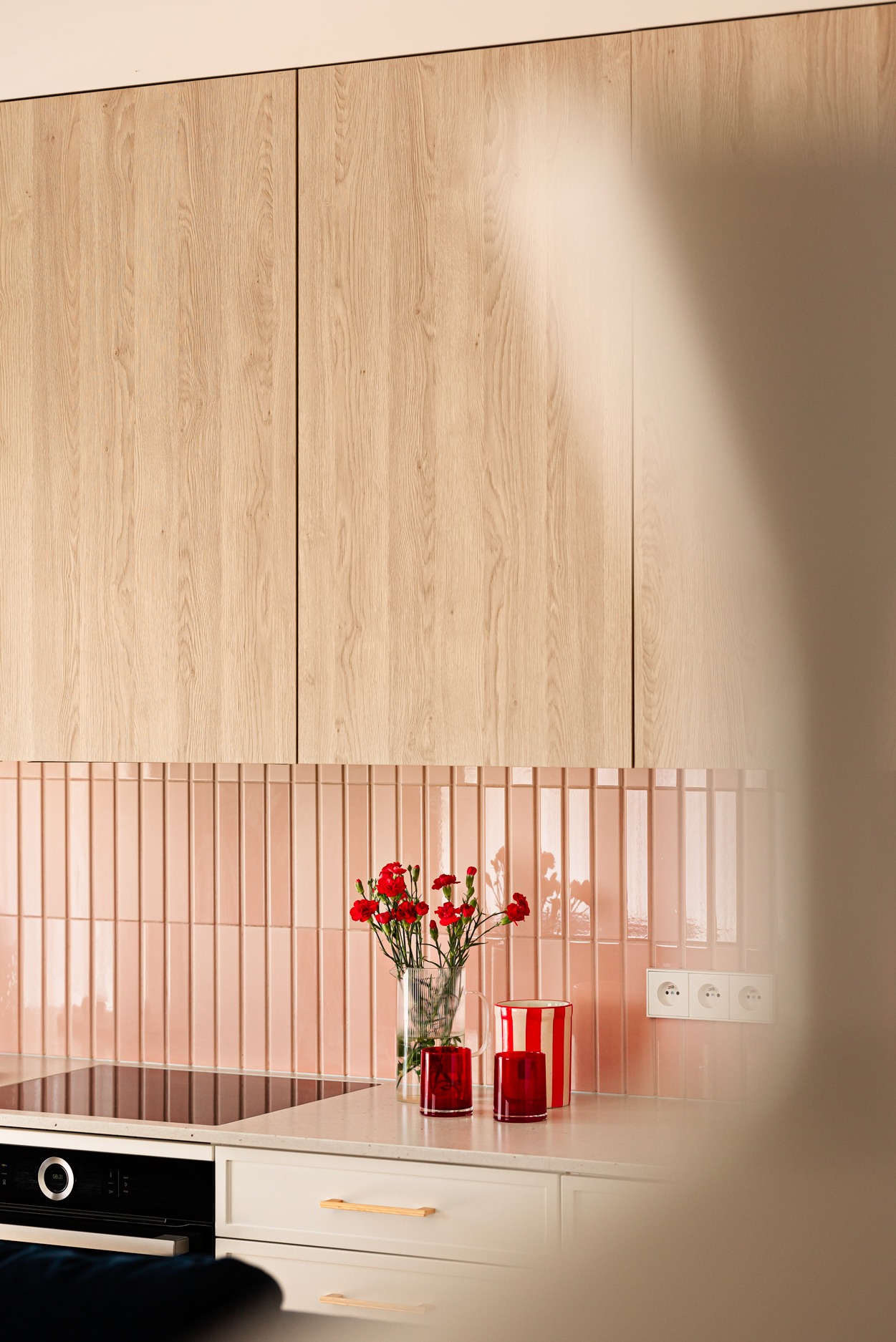 Jasne drewniane szafki kuchenne nad blatem z różowymi, pionowymi płytkami. Na blacie wazon z czerwonymi goździkami i czerwone szklanki. Minimalistyczny design.