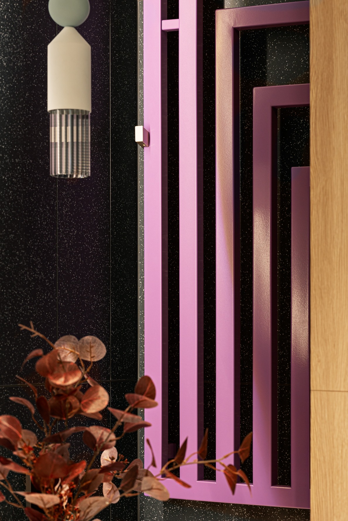 Nowoczesna łazienka: czarne płytki z brokatem, różowy grzejnik dekoracyjny, designerska lampa i brązowe liście w wazonie. Minimalistyczny, ale wyrazisty detal.