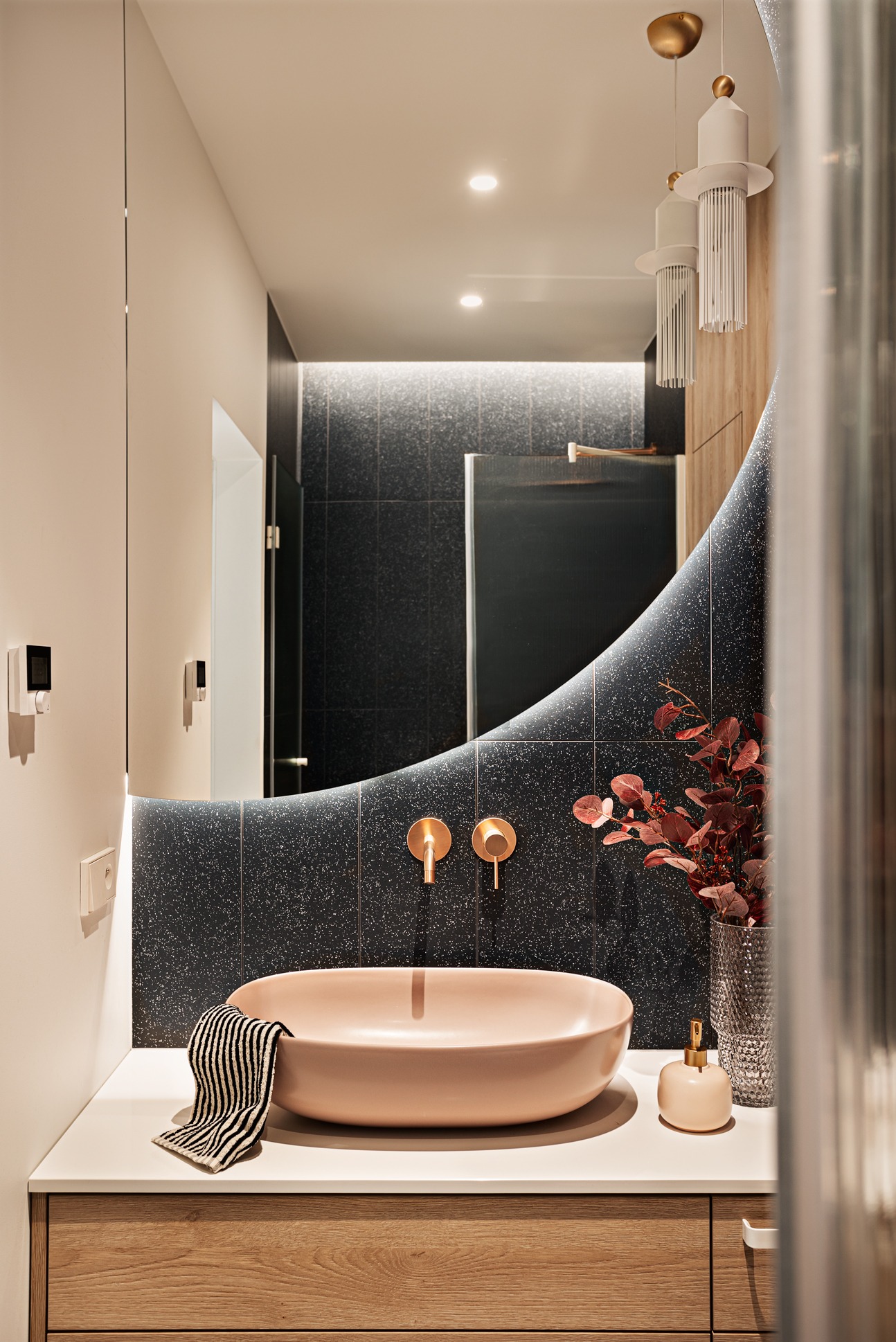 Nowoczesna łazienka z umywalką w odcieniu pudrowego różu, złotą armaturą i lustrem o nieregularnym kształcie. Czarna ściana z mozaiką i drewniane szafki dodają elegancji.