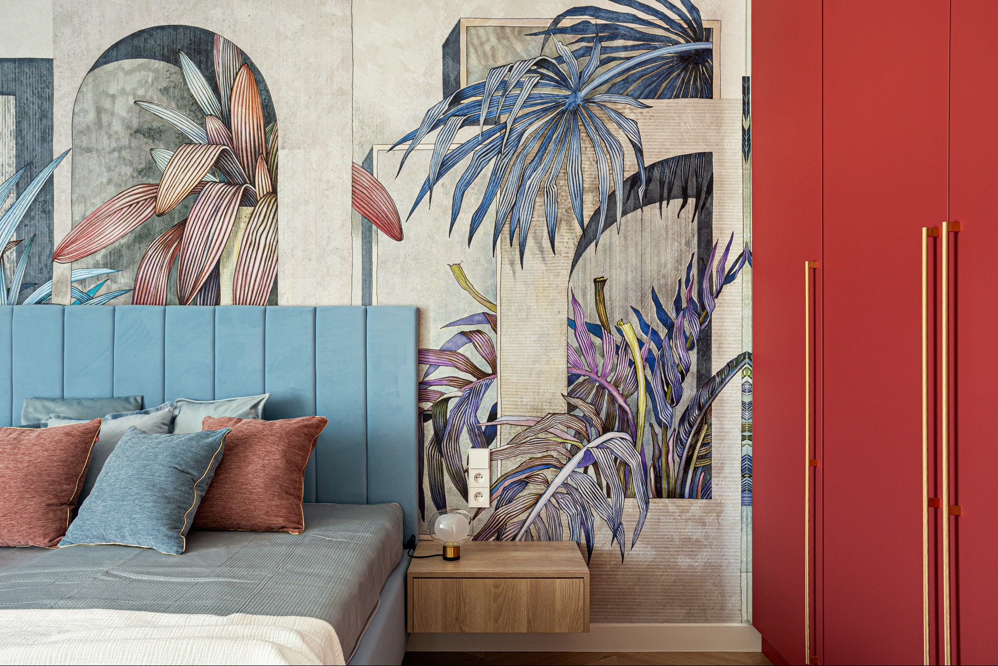 Sypialnia z niebieskim zagłówkiem, wzorzystą tapetą w tropikalne rośliny i czerwonymi szafami. Nowoczesny design z lampką nocną na drewnianym stoliku.