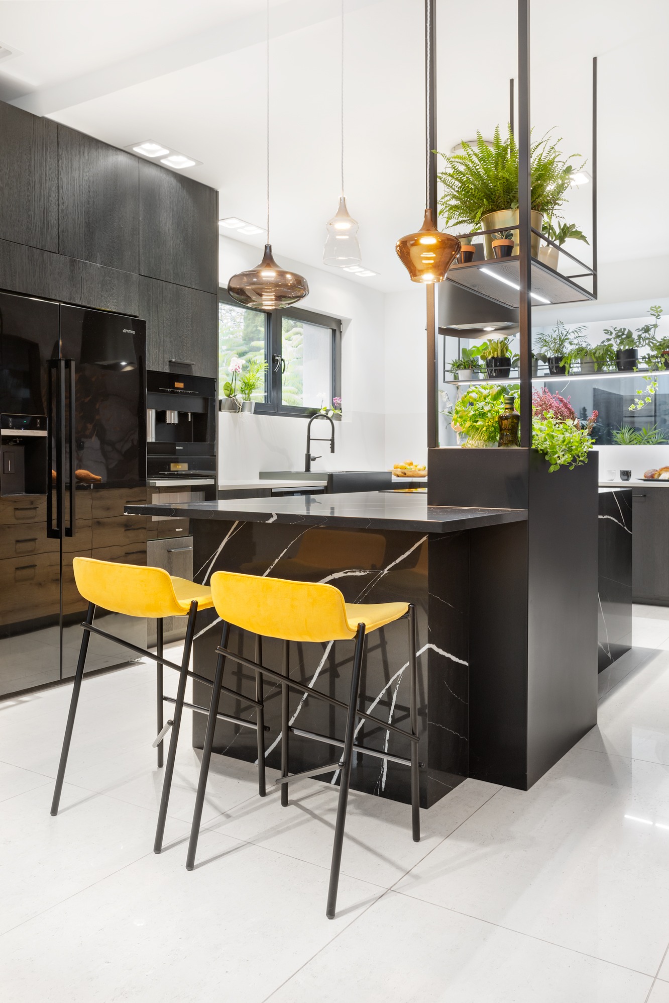 Nowoczesna kuchnia z czarnym marmurem na wyspie i blacie, żółte hokery, wiszące lampy i półki z roślinami. Minimalistyczny design z akcentami zieleni i ciepłego oświetlenia.