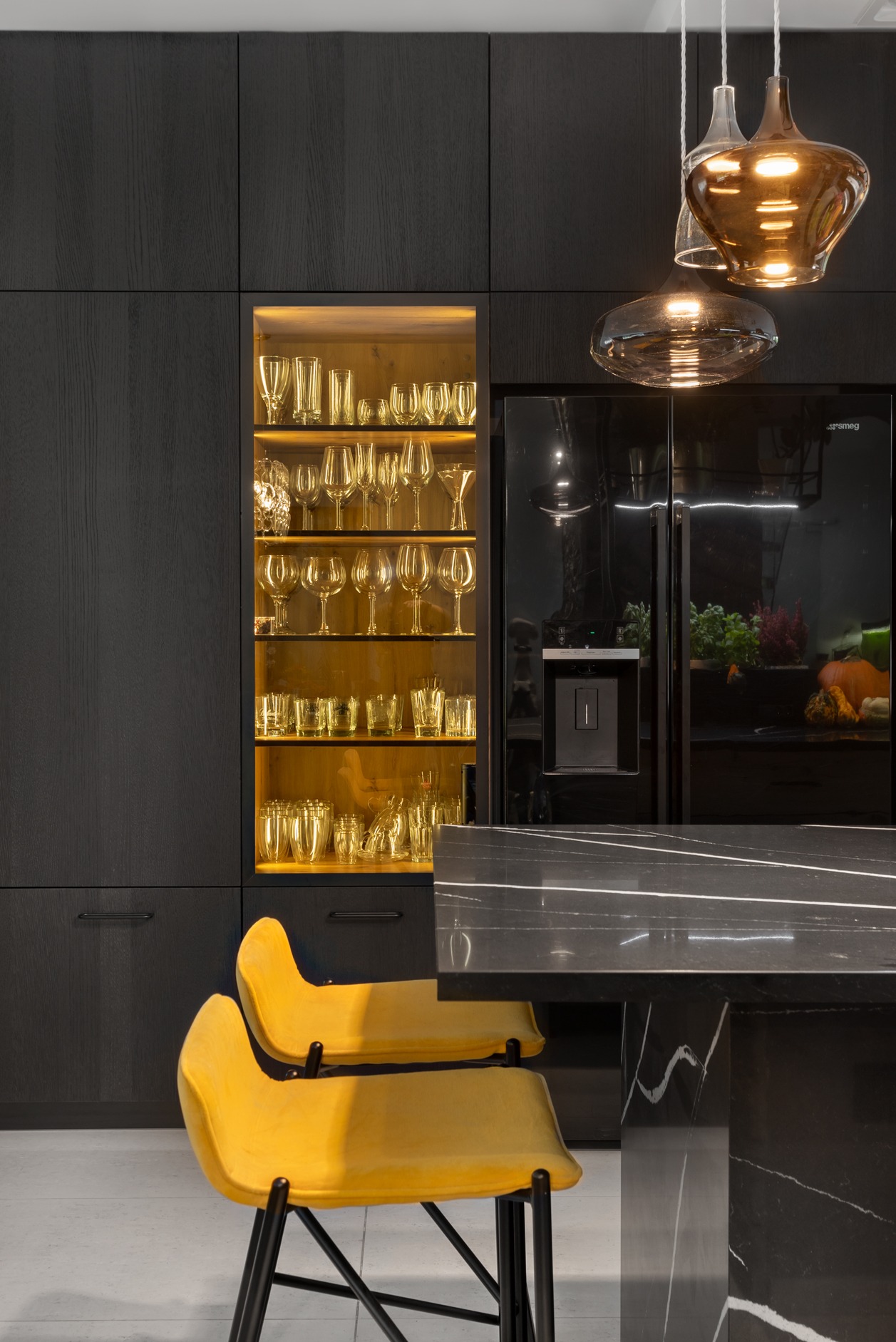 Nowoczesna kuchnia w ciemnych barwach z podświetlaną witryną pełną szkła, czarną lodówką i żółtymi krzesłami barowymi. Lampa wisząca z bursztynowego szkła.