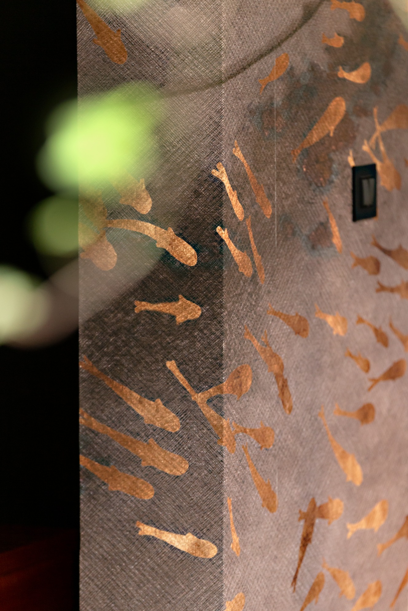 Ściana z dekoracyjnym motywem złotych rybek, oświetlona z boku, z widocznym włącznikiem światła. Nietypowe ujęcie z bliska, z elementami roślinności w tle.