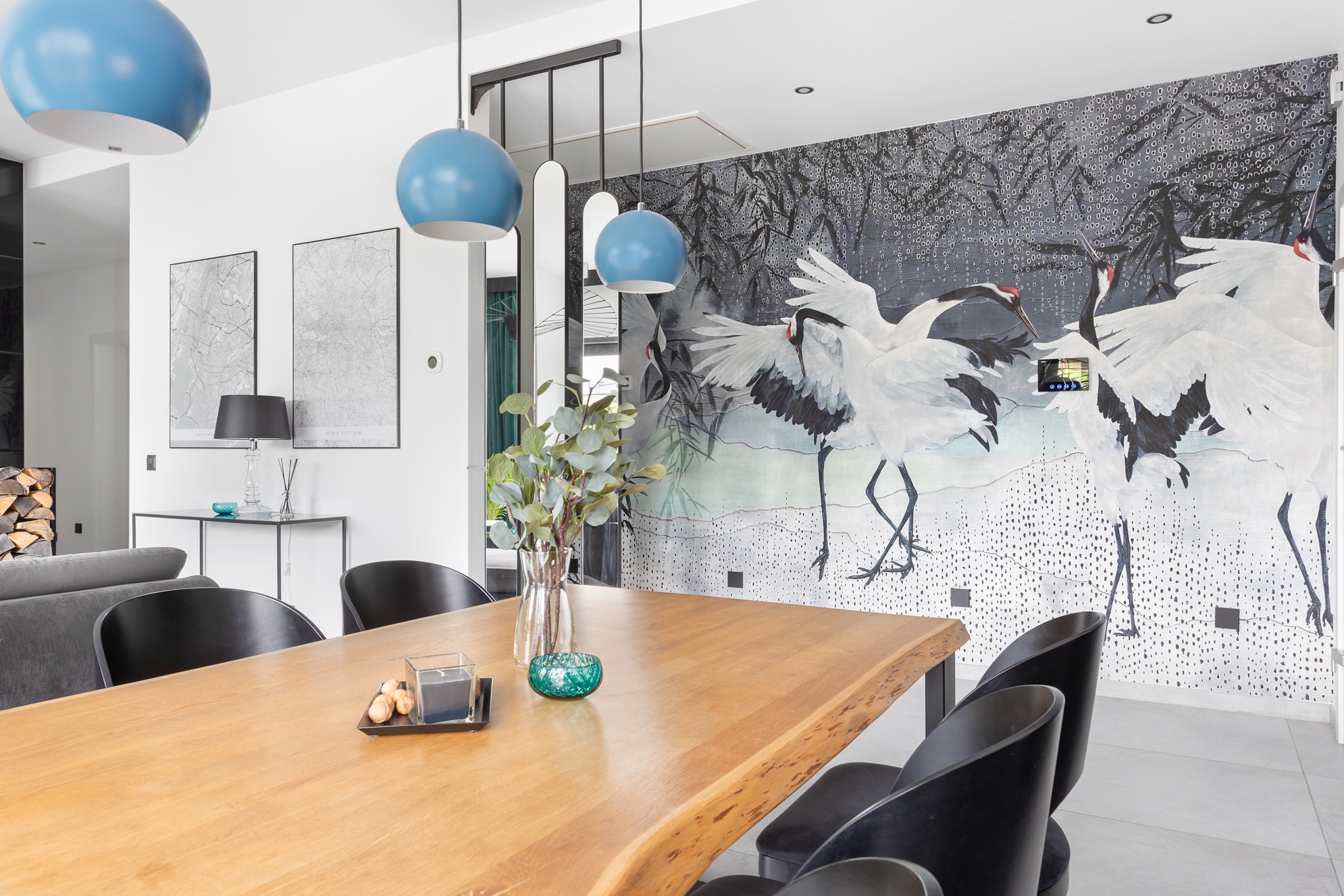 Jasne wnętrze jadalni z drewnianym stołem, czarnymi krzesłami i dekoracyjną tapetą z motywem żurawi. Błękitne lampy wiszące dodają koloru. Fot. Dekorialove Anna Laskowska.