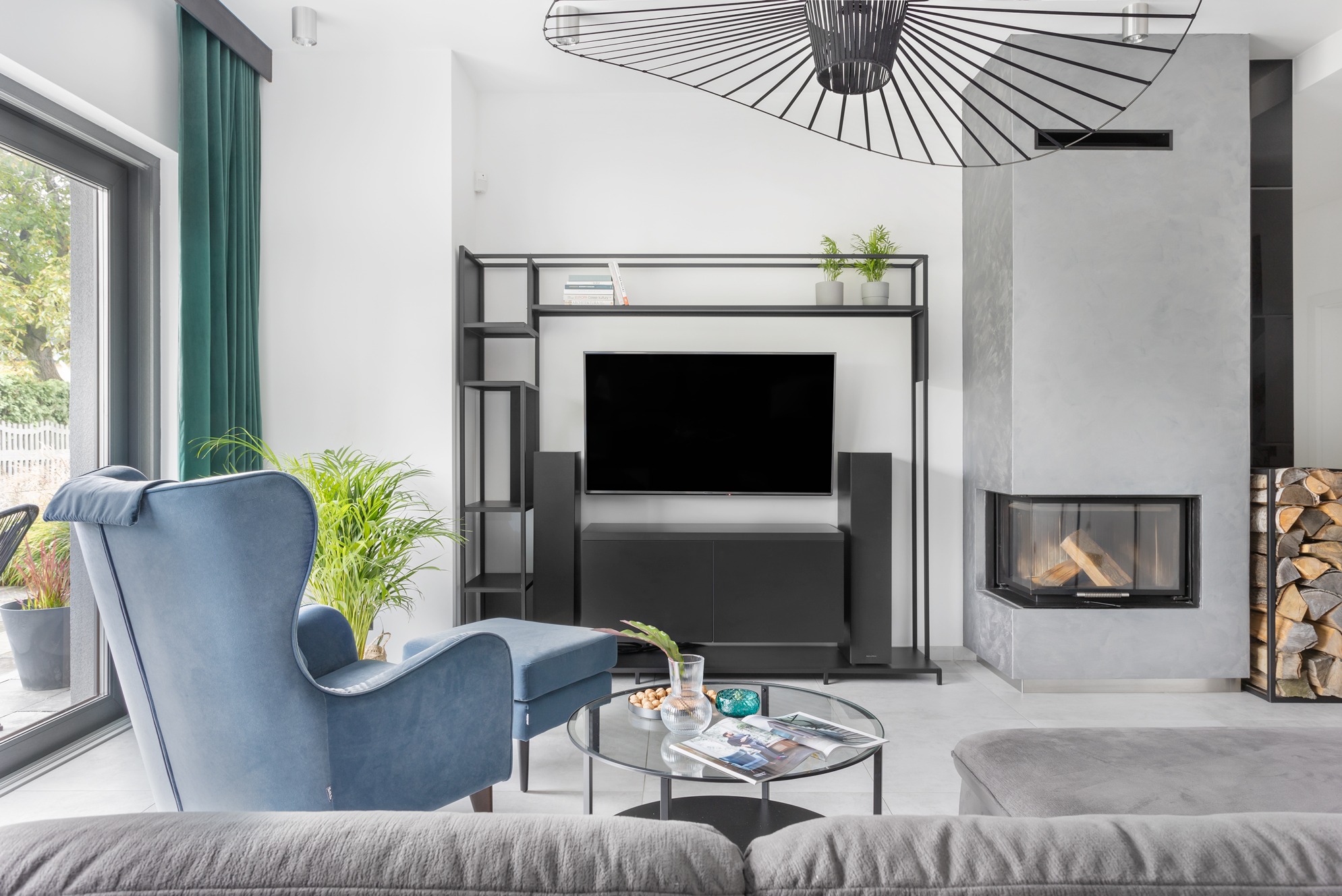 Nowoczesny salon z kominkiem, szarym narożnikiem i błękitnym fotelem uszakiem. Minimalistyczny design, czarne regały i lampa. Widok na zewnątrz przez duże okno.