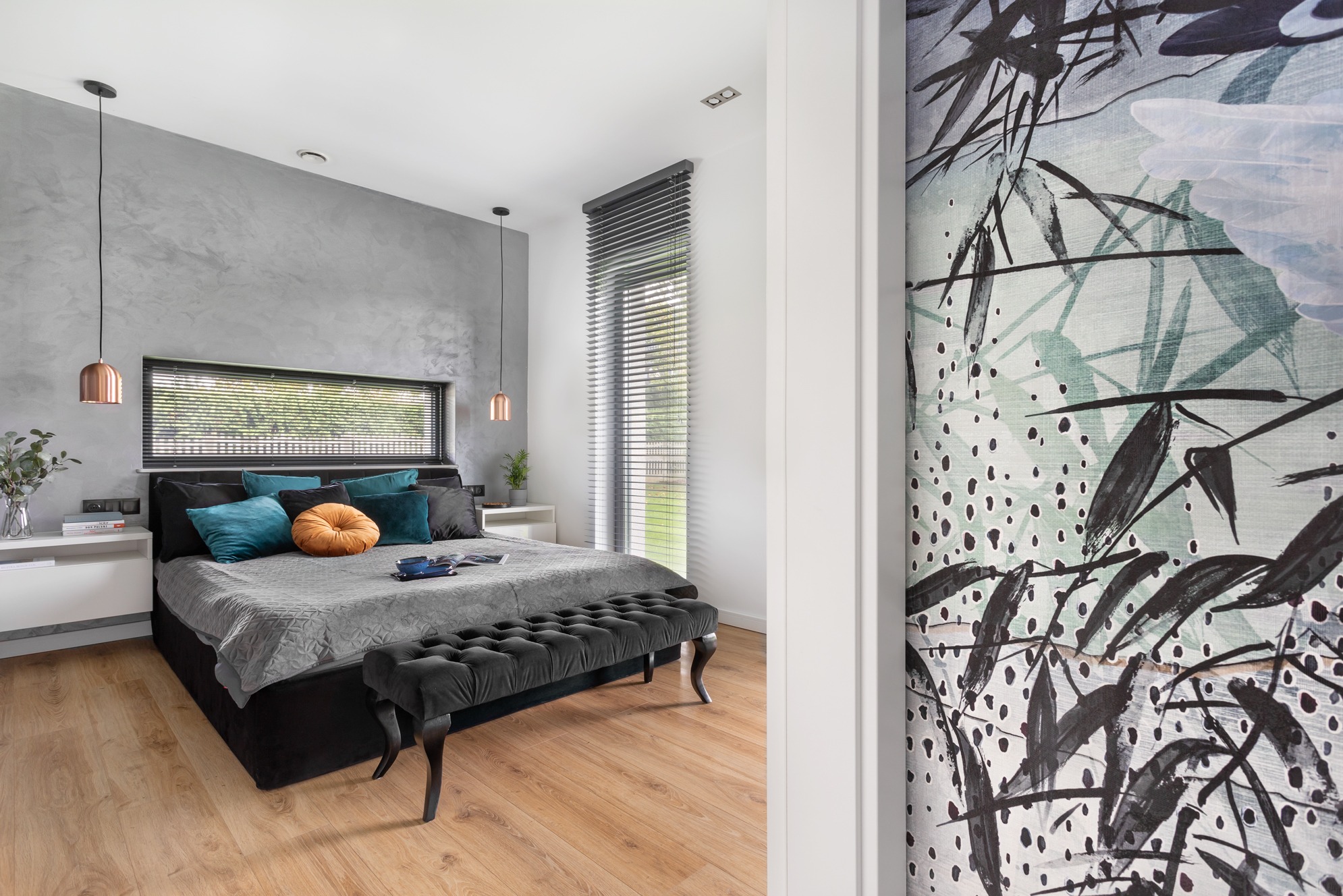 Nowoczesna sypialnia z szarą ścianą, łóżkiem z turkusowymi poduszkami i okrągłą, pomarańczową poduszką. Na podłodze drewniany parkiet, widok na obraz z motywem roślinnym.