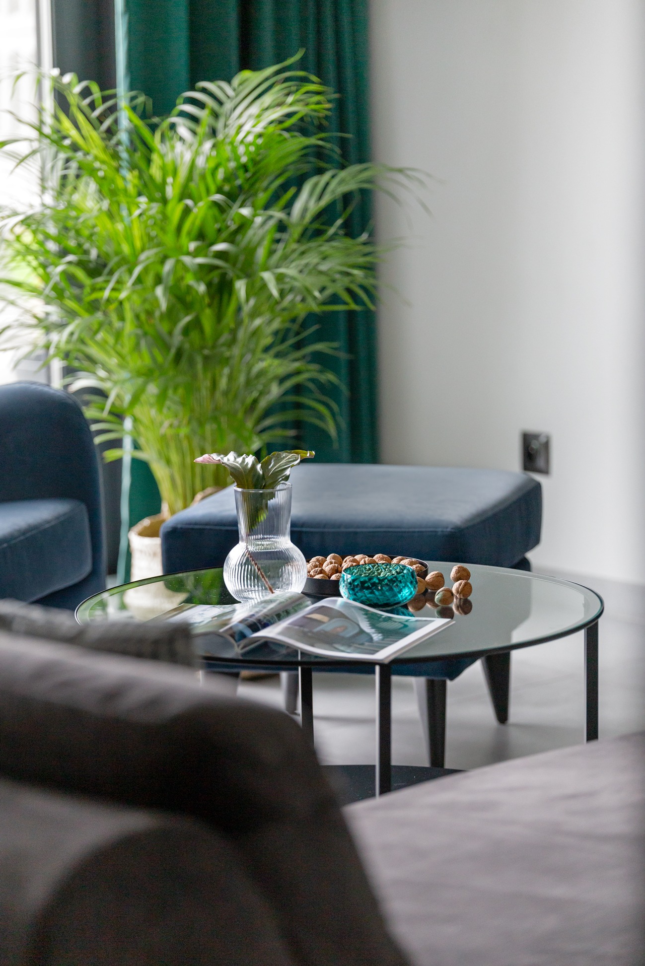 Stylowy salon z okrągłym szklanym stolikiem, na którym leżą magazyny, dekoracyjna misa i wazon z liściem. W tle zielona roślina i zasłony.