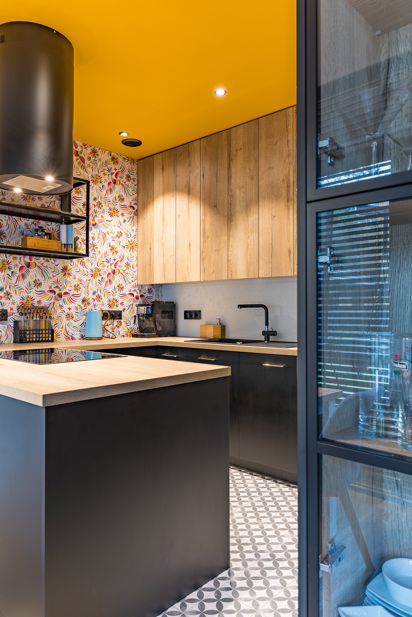 Nowoczesna kuchnia z żółtym sufitem i wzorzystą tapetą. Czarna wyspa z drewnianym blatem, czarne szafki i armatura. W tle przeszklona witryna i geometryczna podłoga.