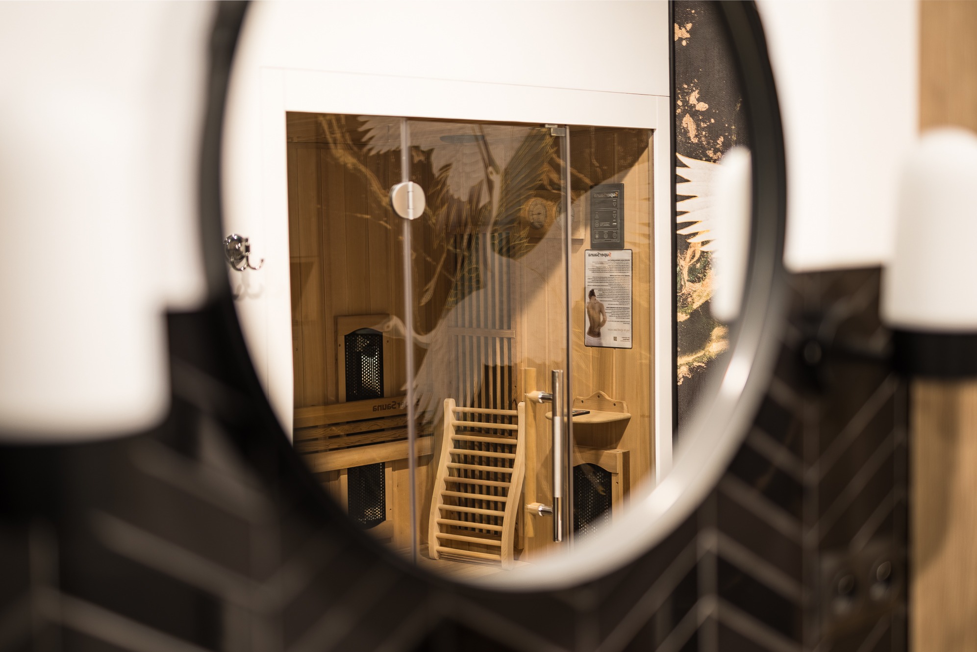 Sauna z drewnianym wyposażeniem, widziana w okrągłym lustrze z czarno-białą mozaiką w tle. Na szklanej ścianie sauny widoczny motyw żurawia.
