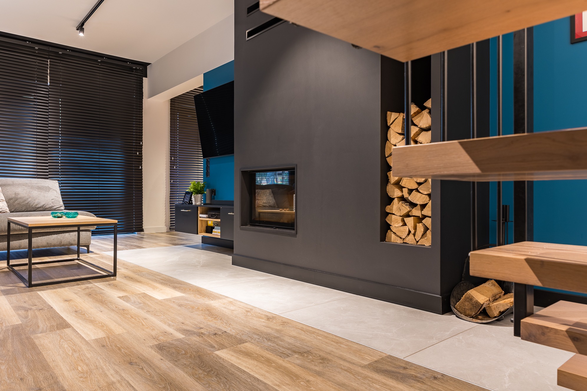 Salon z kominkiem i drewnem opałowym, drewniana podłoga, szare meble, niebieska ściana. Nowoczesne wnętrze z elementami naturalnymi i minimalistycznym designem.