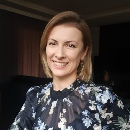 Kamila Kaliszewska-Siwek Psycholog - Uśmiechnięta blondynka w kwiecistej bluzce, z kolczykami, na tle ciemnego wnętrza. Portret kobiety z bliska, z widocznymi detalami makijażu i fryzury.