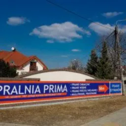 Szyld reklamowy pralni chemicznej 'Pralnia Prima' z listą oferowanych usług (pranie chemiczne, wodne, odzieży wełnianej, maglowanie, prasowanie) na tle błękitnego nieba i budynku z czerwoną dachówką.