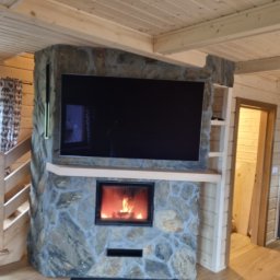 Łukasz Golec - Salon w stylu rustykalnym z kamiennym kominkiem, w którym umieszczony jest telewizor. Drewniane wykończenie sufitu i ścian, podłoga z paneli. W kominku płonie ogień.