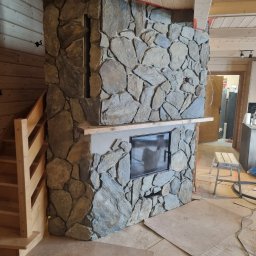 Łukasz Golec - Kamienna obudowa kominka z wkładem kominkowym, drewniane schody i sufit, zabezpieczona podłoga kartonem, wnętrze w trakcie wykończenia.