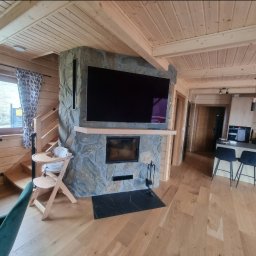 Łukasz Golec - Drewniany salon z kamiennym kominkiem, na którym wisi telewizor. Widoczny drewniany sufit, podłoga i schody z dziecięcym krzesełkiem. Czarny marmurowy element pod kominkiem.