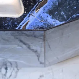 Fragment wykończonej łazienki z umywalką, blatem z czarnego marmuru z niebieskimi żyłami i białymi płytkami podłogowymi z szarymi wzorami.