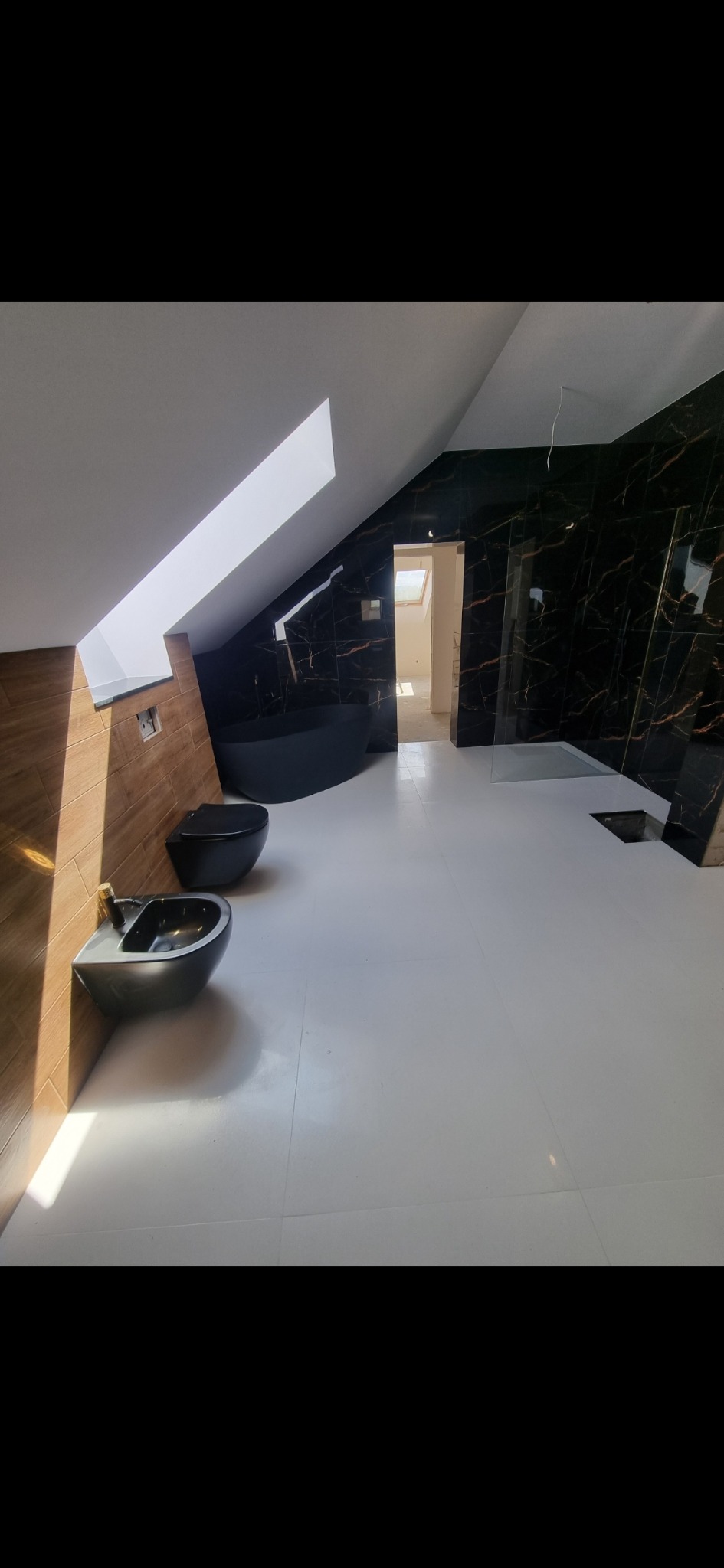 Nowoczesna łazienka na poddaszu z czarną armaturą: wolnostojąca wanna, bidet i toaleta. Ściany wyłożone czarnymi płytkami z marmurkowym wzorem, podłoga z jasnych płytek. Światło dzienne wpada przez...