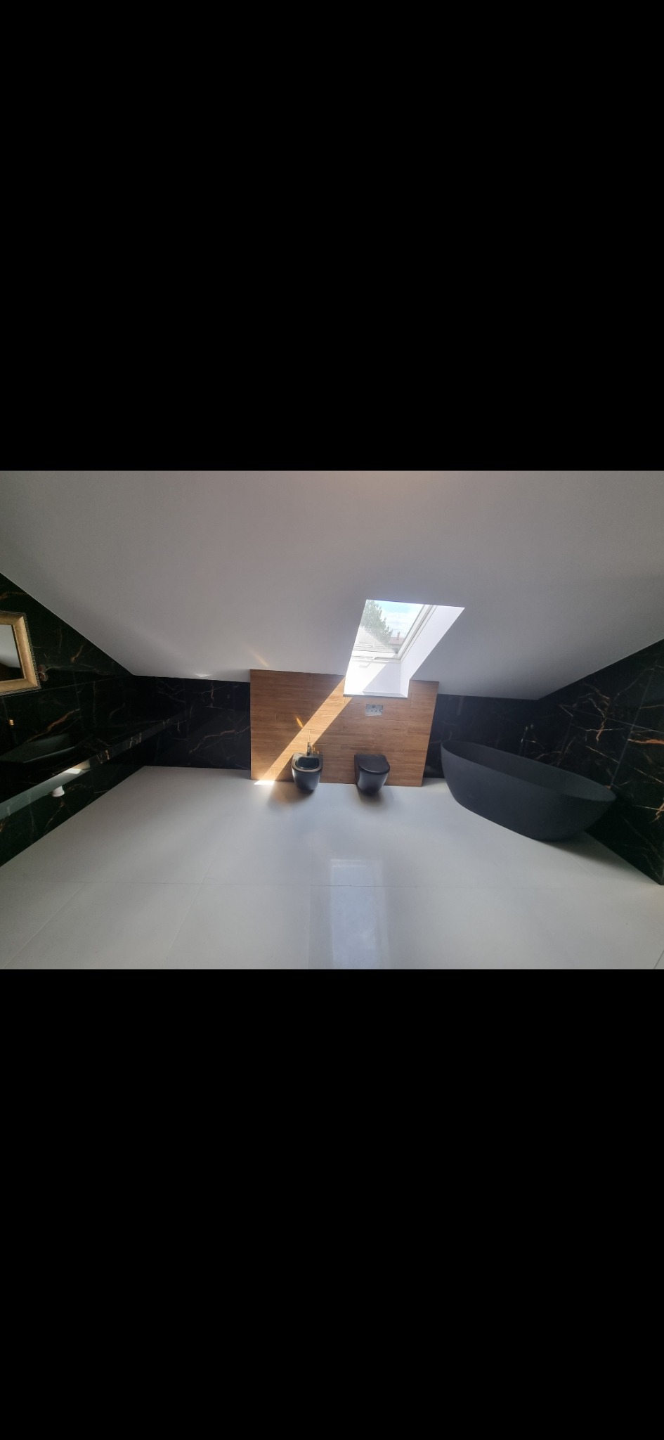 Nowoczesna łazienka na poddaszu z czarnymi płytkami imitującymi marmur, białymi płytkami podłogowymi, drewnianą ścianą, czarną wanną wolnostojącą i oknem dachowym wpuszczającym jasne światło.