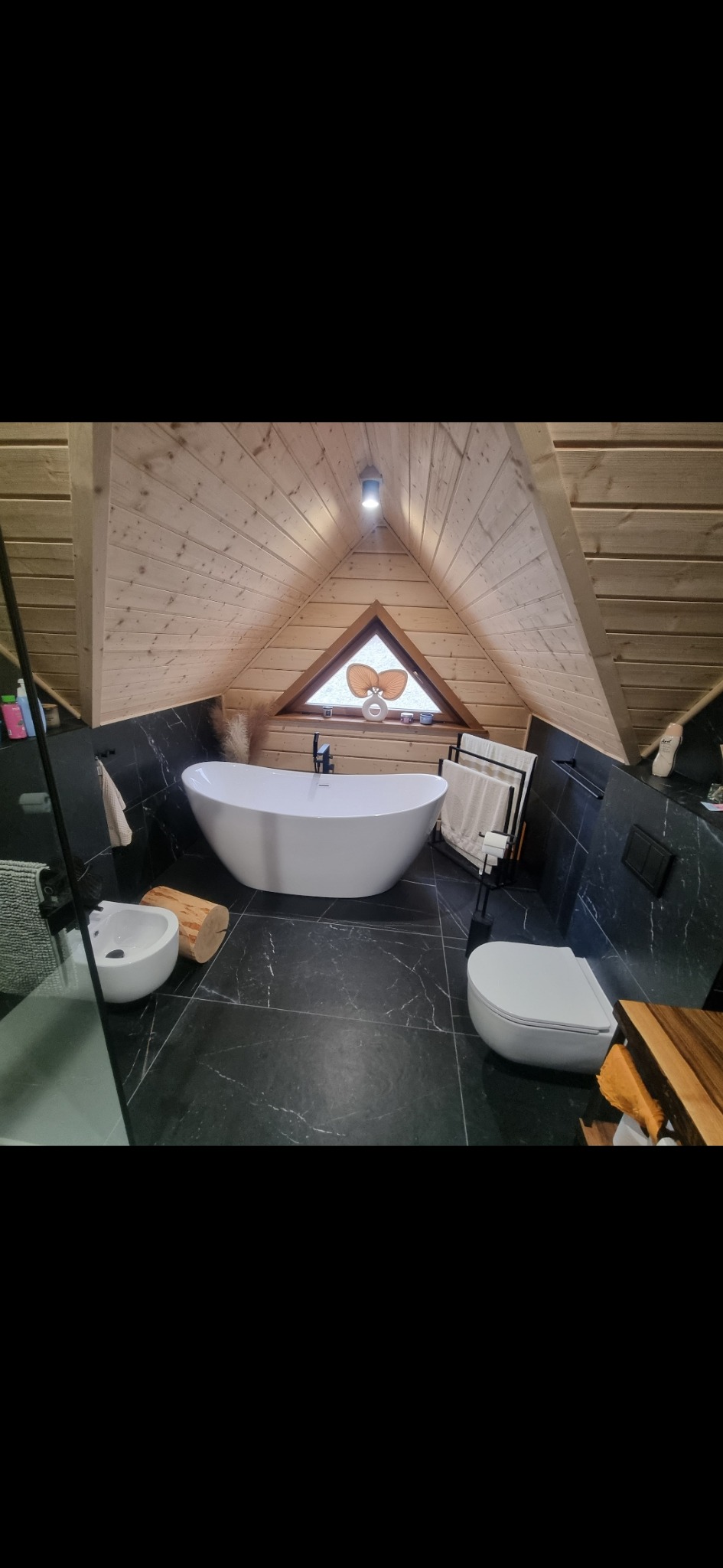 Nowoczesna łazienka na poddaszu z drewnianą boazerią, czarnymi płytkami imitującymi marmur, wolnostojącą białą wanną i triangularnym oknem z dekoracją.