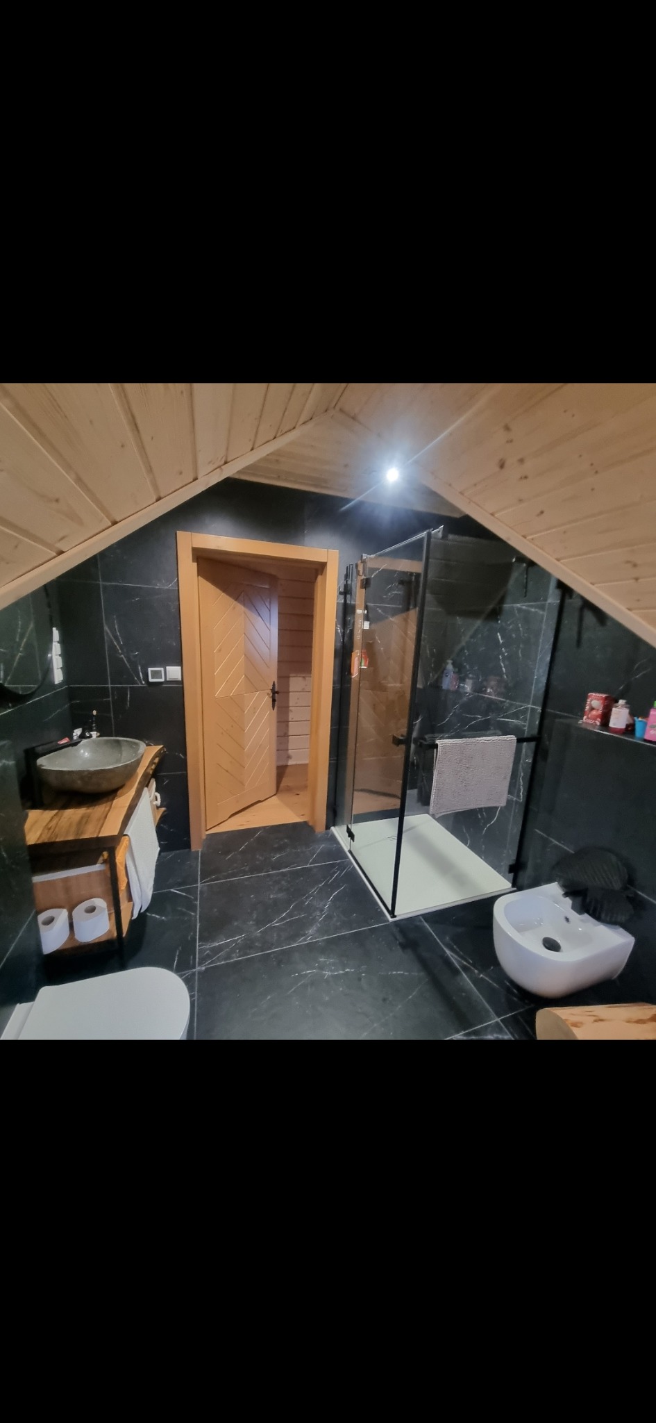 Nowoczesna łazienka na poddaszu z czarnymi marmurowymi płytkami, drewnianym sufitem i drzwiami z motywem jodełki, wyposażona w kabinę prysznicową typu walk-in, umywalkę z kamienia i bidet.