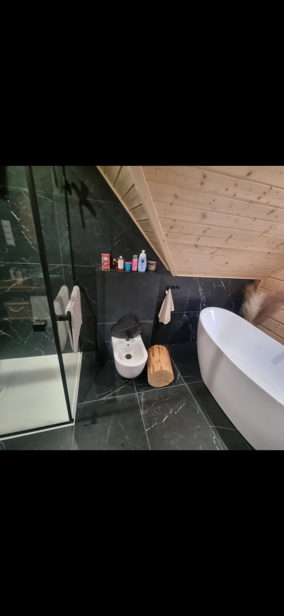 Nowoczesna łazienka na poddaszu z czarnymi marmurowymi płytkami, białą wanną wolnostojącą, bidetem, kabiną prysznicową z czarnymi ramami i drewnianym pieńkiem jako elementem dekoracyjnym.