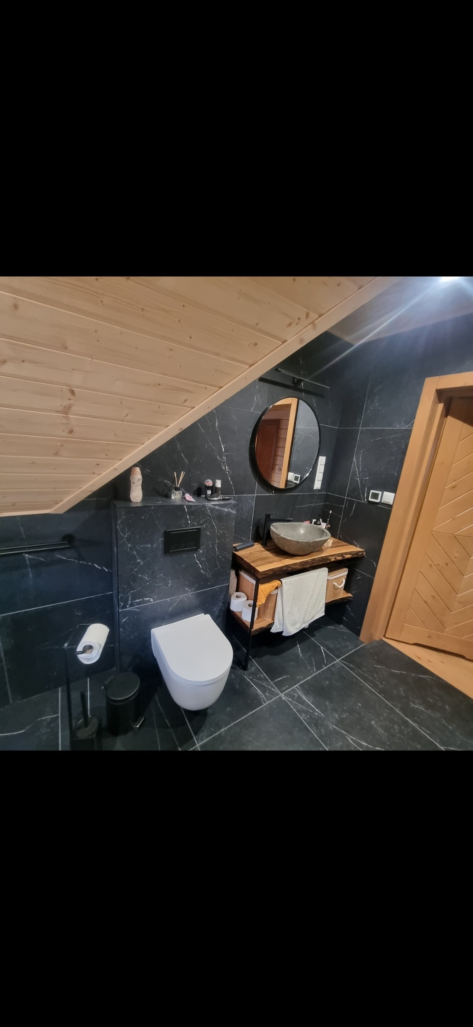 Nowoczesna łazienka na poddaszu z czarnymi płytkami imitującymi marmur, drewnianym sufitem i umywalką z kamienia na drewnianym blacie.